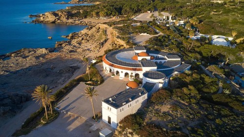 Drone shot, morning light, A round building on the rocky Mediterranean coast, surrounded by pine