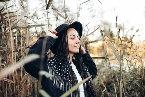 A woman stands peacefully in a field during sunset, surrounded by tall grasses. She wears a hat and