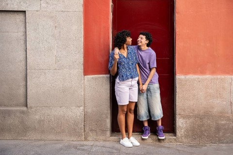 A joyful multiethnic LGBTQ+ couple stands together outdoors, holding hands in a warm embrace,