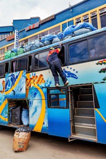 A person climbs onto the roof of a bus to load luggage in an urban setting, bus trip from La Paz to