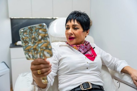 Mature woman in a bright aesthetic clinic chair smiling as she checks her refreshed face in a