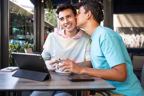 A joyful gay couple shares coffee and a tender moment at an outdoor cafe. One leans in for a kiss,