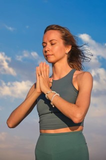 A woman in yoga holds Namaskar Mudra while standing on the beach at sunset, focusing on meditation