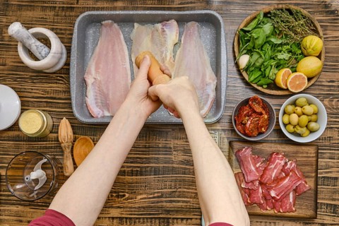 Hands sprinkle fish fillet with salt while surrounded by fresh herbs, olives, and sliced meats