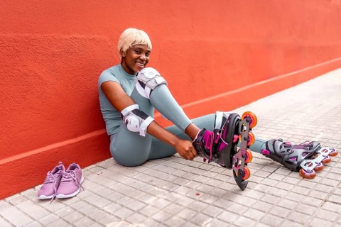 Happy african sportive woman putting on inline skates sitting in the street leaning on red facade