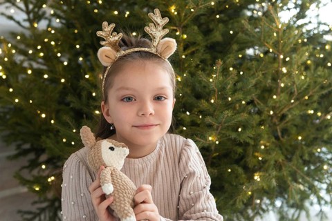 A young girl with blue eyes wearing reindeer antlers smiles gently while holding a plush toy,