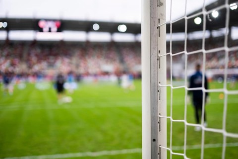 Close-up of a white soccer goalpost and net on a green grass pitch with blurred players training in