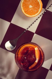 A drink filled with ice cubes and orange slices is placed on a checkered table. A metal spoon lies