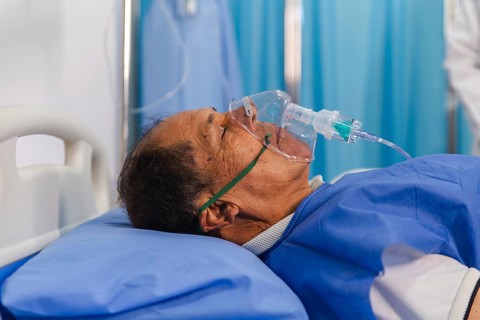 An elderly patient lying in a hospital bed receiving oxygen therapy through a mask. The scene