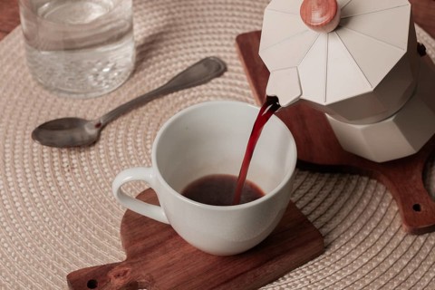 A coffee maker pours steaming coffee into a white cup placed on a wooden mat. Nearby, a glass of