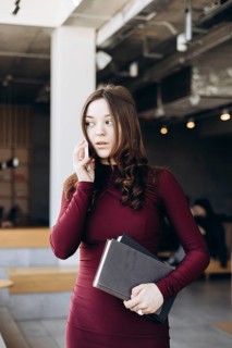 A young female student stands indoors, balancing books while talking on her phone. Her expression