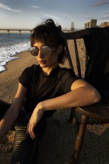 A stylish woman wearing unique sunglasses relaxes on a chair at a sandy beach. The serene ocean