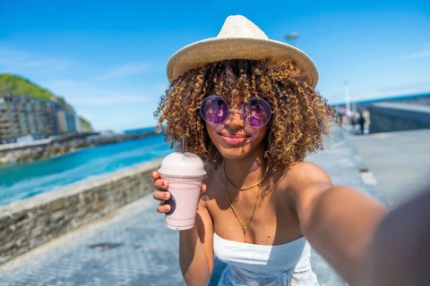 Cool woman with curly hair and hat taking a selfie drinking milkshake next to the sea