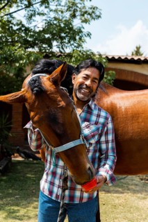 A man in a plaid shirt shares a joyful moment with his horse in a sunny garden. The horse nuzzles