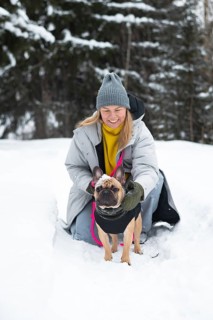 A woman wearing a grey coat and beanie kneels in the snow, smiling at her French bulldog. Snowy