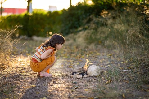 A young girl crouches down and gently blows on a small fire, watching the flames with fascination.