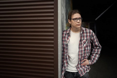 Teenage boy with long hair and glasses leaning against a wall outdoors. He wears a plaid shirt over