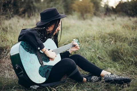 A woman with long braids plays an acoustic guitar while sitting in a grassy field at sunset. She