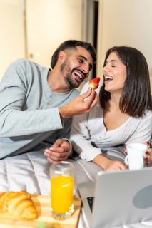 Young man offering a strawberry to his smiling girlfriend during a romantic breakfast in bed, with