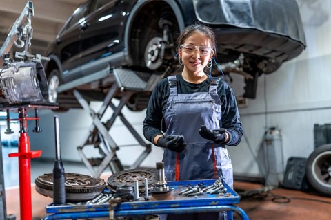 Confident young female mechanic wearing gloves and protective glasses, working on car parts in a