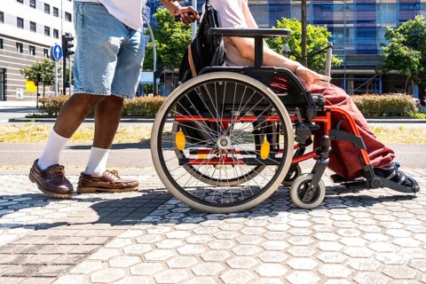 Caregiver pushing a senior person in a wheelchair along an accessible city sidewalk, providing