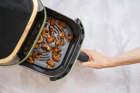 A hand holds the basket of an air fryer that contains cooked champignon mushrooms. The mushrooms
