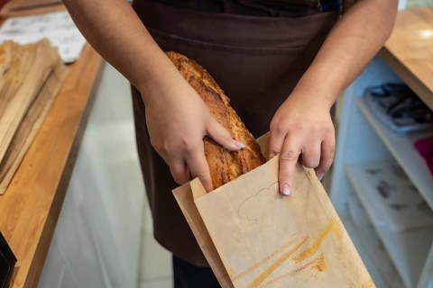 Baker's hands carefully placing a fresh, crusty artisan bread loaf into a brown paper bag,