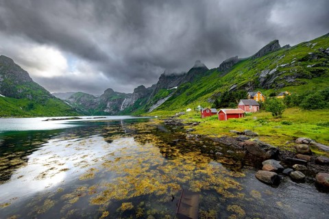 Red wooden houses on the shore in green fjord landscape with mountains and dramatic clouds,