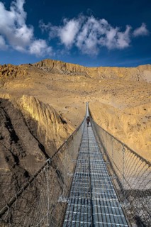 A man walks over a very long suspension bridge made of steel cables across a river valley, mountain