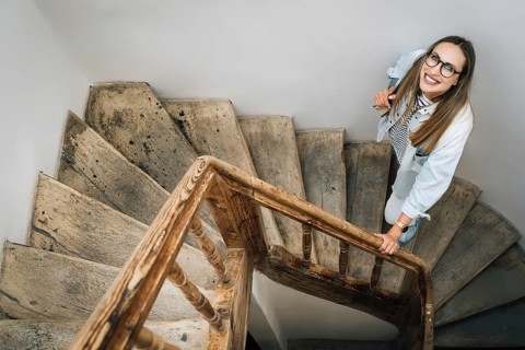 A woman with glasses smiling looks up climbing an old wooden staircase, A smiling woman looks up