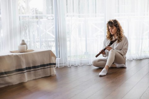 A woman sits on a wooden floor holding a flute, with natural light streaming in