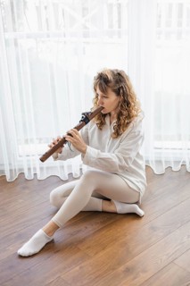 A seated woman playing a flute in a bright room with a wooden floor