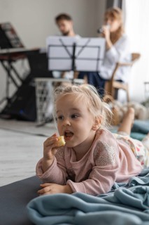 A young Caucasian girl lies on a blanket, eating a snack. In the background, a man plays keyboard