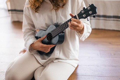 Person kneels with a ukulele on a wooden floor, wearing a casual attire