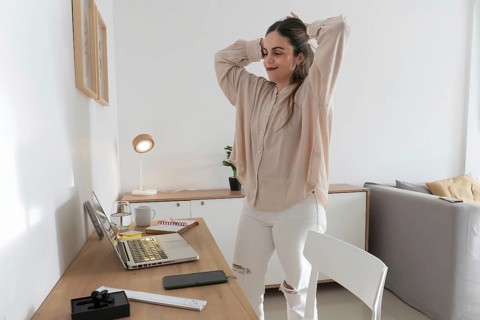 Woman taking a break from work, stretching arms and tying her hair up, standing beside desk with