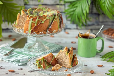 A sliced cake and pieces of cake with green icing next to a cup of coffee and almonds