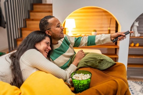 Happy smiling multiracial couple cuddled on a cozy sofa, sharing popcorn and a remote while