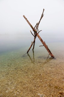 On a foggy morning, a wooden figure made of three branches stands in Lake Almsee. Austria