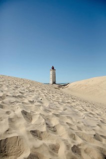 A lighthouse stands majestically surrounded by sand dunes under clear blue skies, lighthouse in the