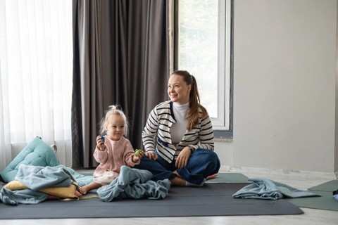 A young Caucasian woman with long brown hair sits on a mat with a toddler girl. The girl has blonde