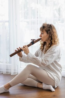 A woman in a white shirt playing a wooden flute while seated on a wooden floor