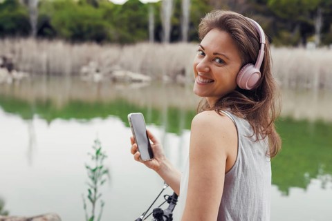 A young Caucasian woman with brown hair wearing headphones stands by a lake, checking her