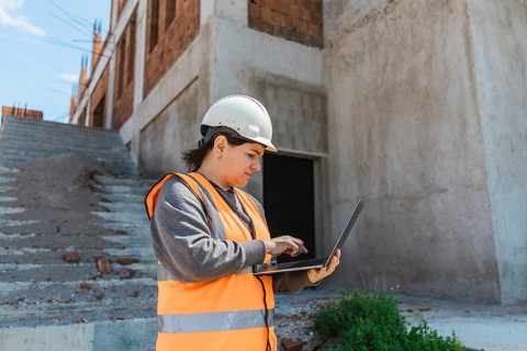 Woman in a hard hat and safety vest uses a laptop on a construction site. The background features