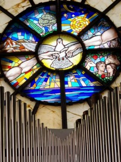Artistic church window with a dove surrounded by colorful stained glass depictions of organ pipes,