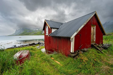Red wooden houses in green mountain landscape with dramatic clouds, Vindstad, Reinefjord, Lofoten,