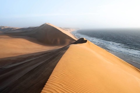 Sand dunes with wave patterns and sea in evening light, wind blowing sand across the dunes,