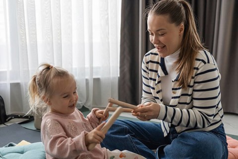 A young Caucasian woman and a little girl play with wooden music sticks at home. They are smiling