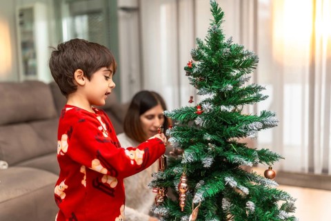 Young boy in red pajamas decorating a small christmas tree with ornaments while his mother sits in