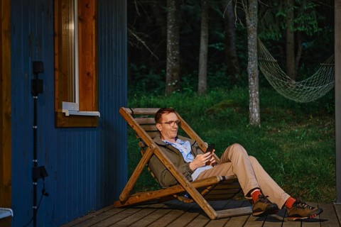 Middle-aged man sits on wooden chair outside wooden cabin in forest. He uses smartphone and enjoys