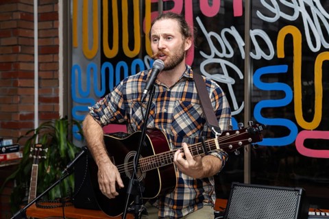 A young Caucasian man with a beard sings into a microphone while playing an acoustic guitar.
